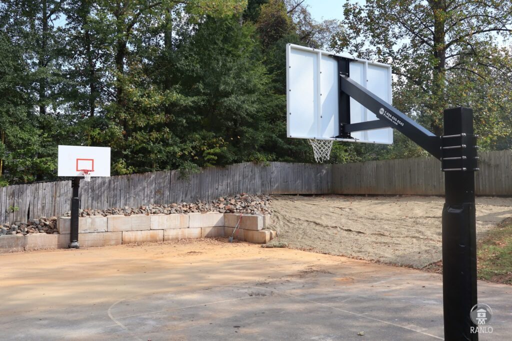 Gardin Park basketball court with a newly installed black basketball hoop structure in the foreground, a second new hoop in the background, and a stone retaining wall behind the court.