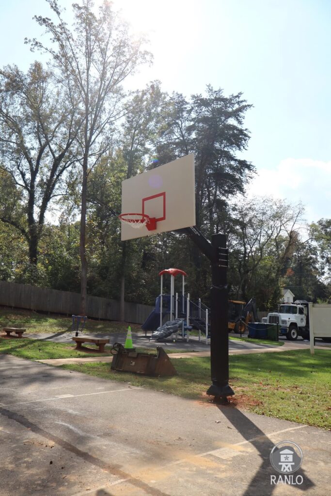 A new black basketball hoop and backboard at Gardin Park, with the park playground and Public Works equipment visible in the background.
